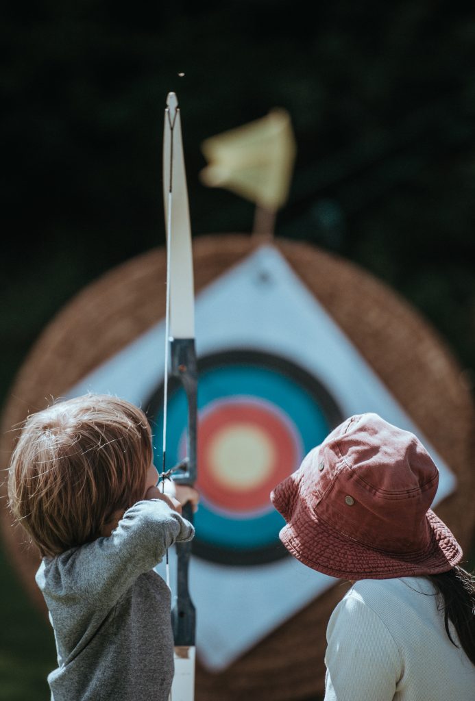 Boy and girl standing facing archery target. Boy has arrow drawn on bow.